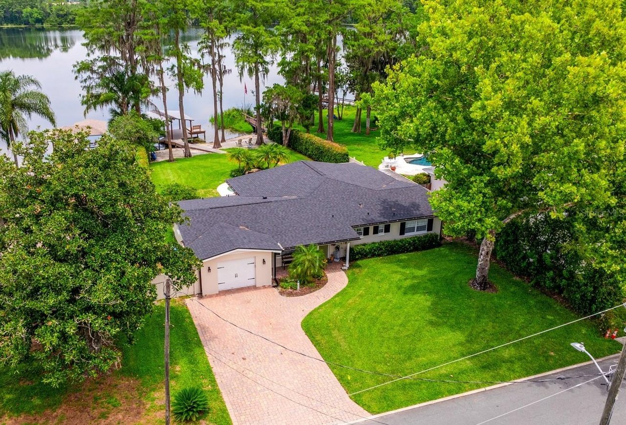 Aerial view of a lakeside home with a curved brick driveway, pool, and wooded shoreline.