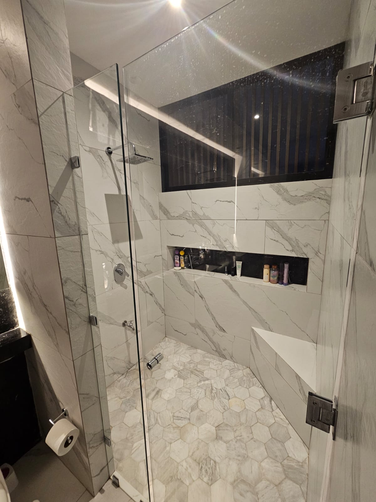 Walk-in shower with marble-look tile, a glass enclosure, recessed toiletry shelf, and built-in bench.