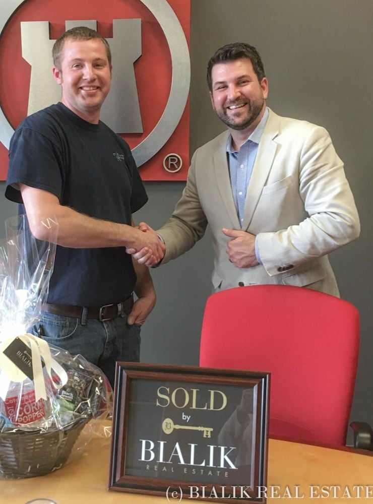 Two men shake hands beside a framed SOLD sign and closing gift basket.