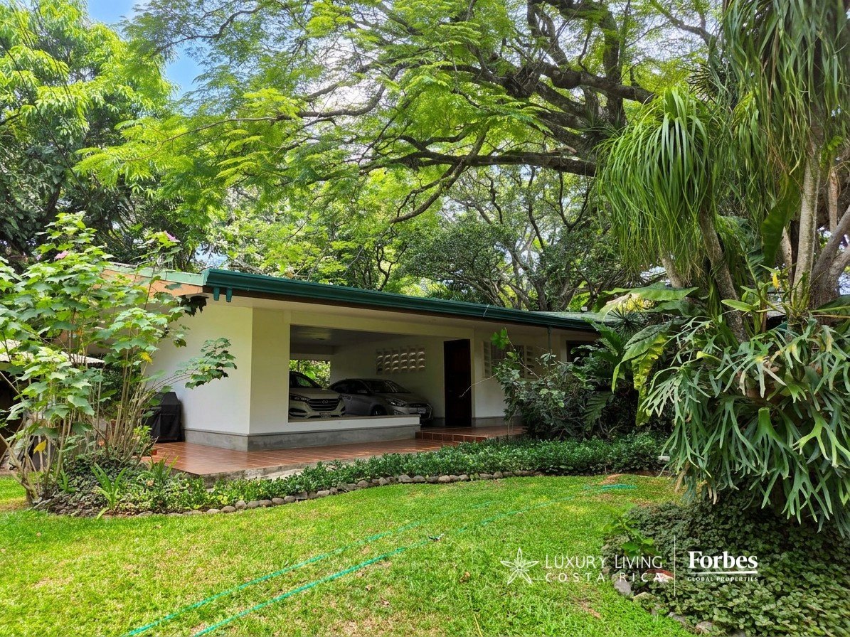 Single-level villa and carport beneath dense tropical trees, with a broad lawn.