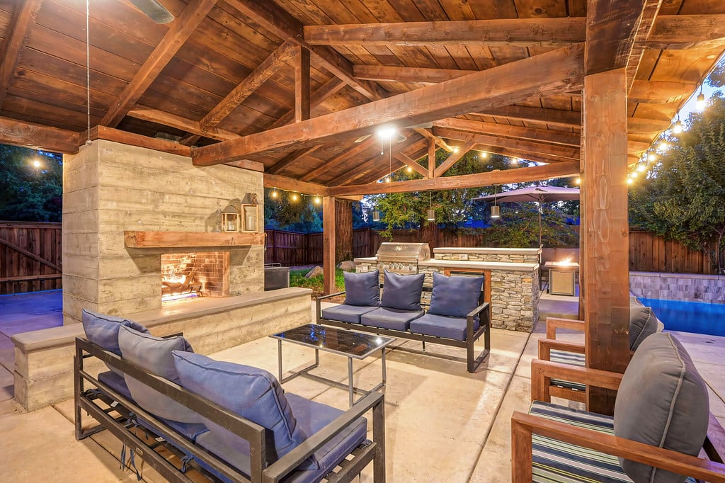 Covered patio with timber beams, a stone fireplace, lounge seating, and a pool at dusk.