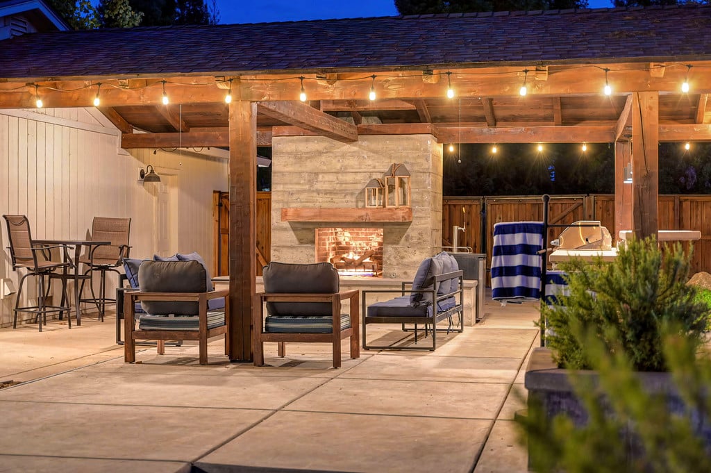Covered patio with a stone fireplace, lounge seating, and warm string lights at dusk.