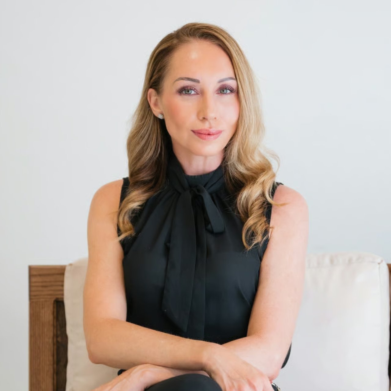 Woman with long blonde hair seated in a light chair, wearing a black sleeveless blouse.