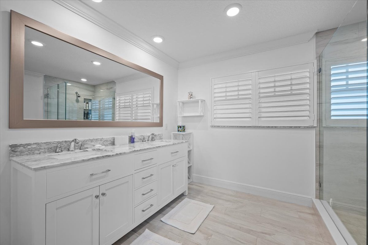 Bathroom with a long white vanity, large framed mirror, shuttered windows, and glass-enclosed shower.