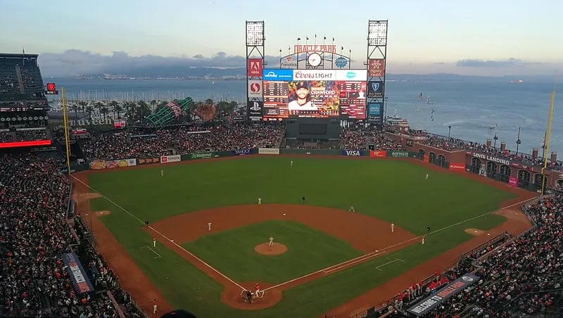 High-angle wide shot of Oracle Park stadium during a San Francisco Giants game, showing the green baseball field, the massive scoreboard, and the scenic San Francisco Bay with McCovey Cove in the background.