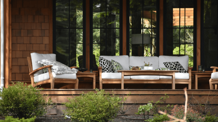 Wooden porch with white-cushioned seating, patterned pillows, and large black-framed windows.