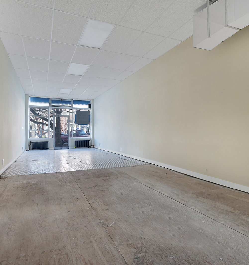 Vacant storefront interior with bare flooring, fluorescent ceiling lights, and large front windows.