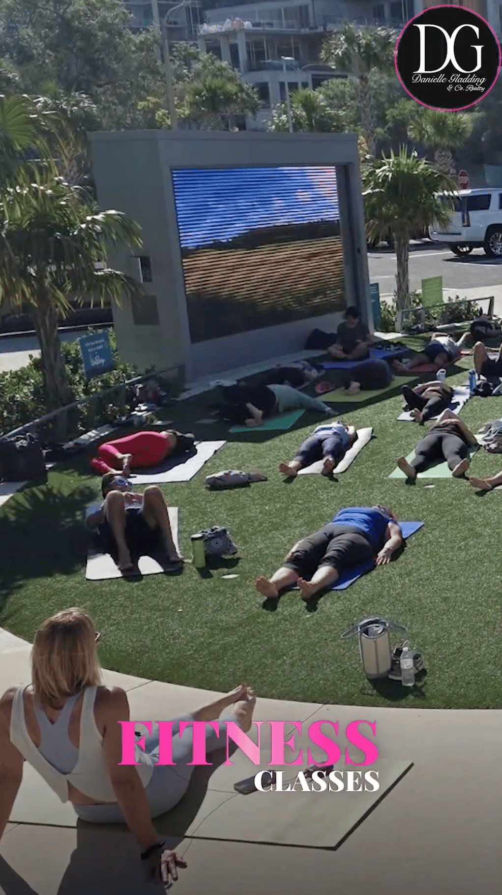 Outdoor fitness class on a park lawn, with participants exercising on mats beside a large digital display.
