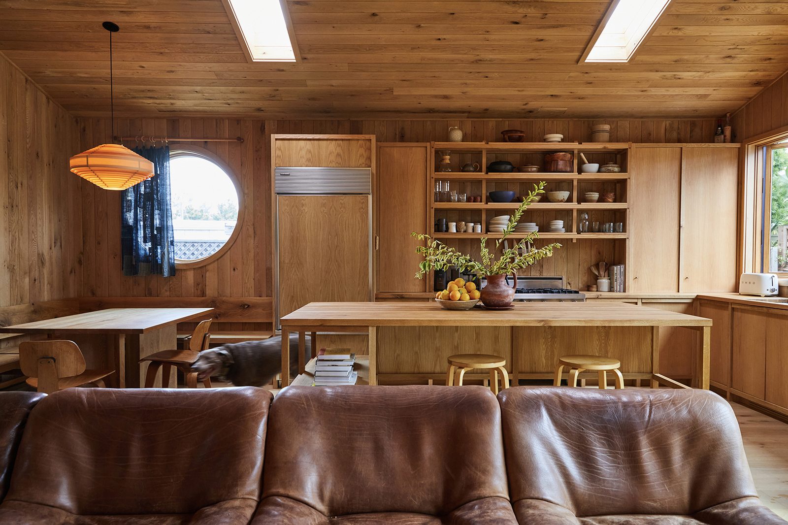 An open oak-paneled kitchen and living room with a vintage De Sede leather sofa, a round window, and built-in shelving.