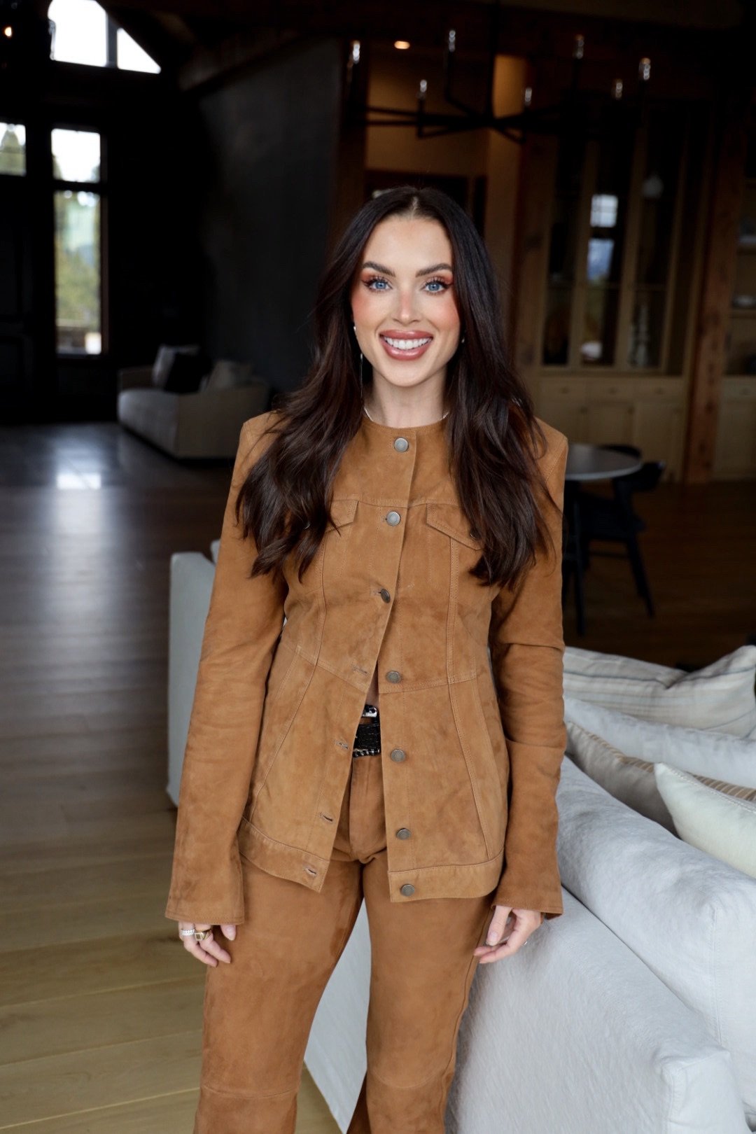 Woman in a tan suede suit standing beside a light-colored sofa in a modern living room.