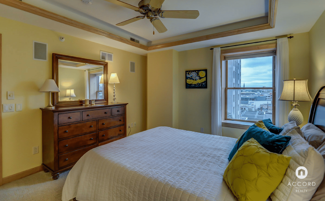 Furnished condo bedroom with a large bed, wooden dresser, and a city view through the window.