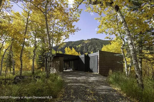 Modern dark-sided home at the end of a gravel drive, framed by golden aspens with a mountain behind it.