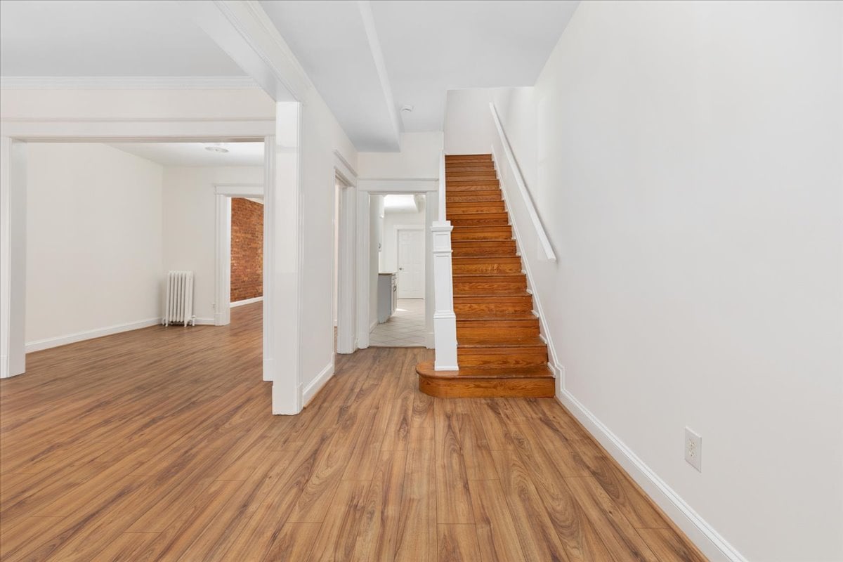 Vacant hallway with warm wood floors and a wooden staircase beside an open doorway.