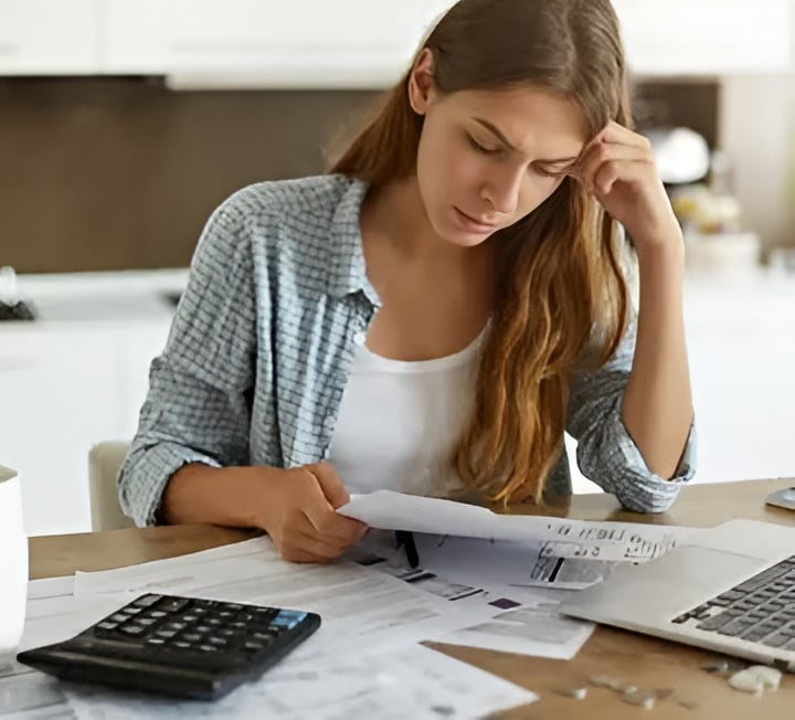Woman reviewing paperwork at a kitchen table beside a calculator and laptop.