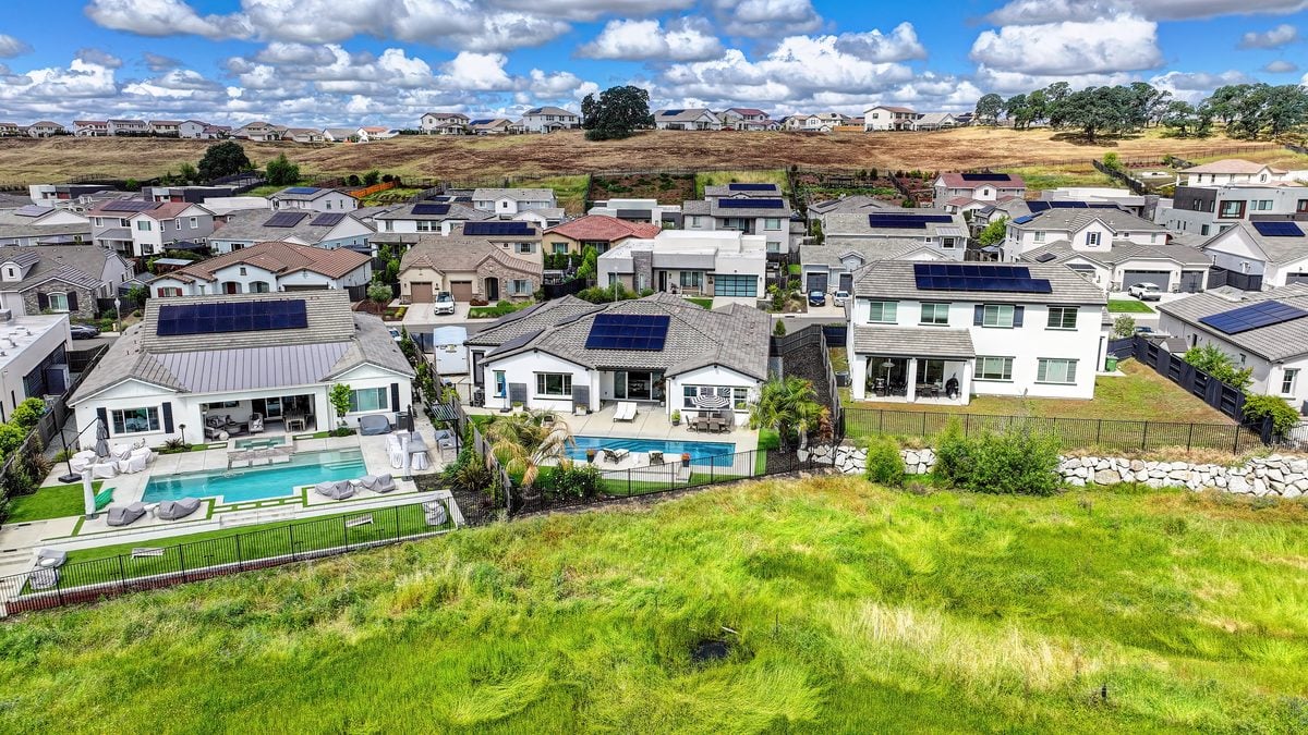 Aerial view of newer single-story and two-story homes with solar panels and backyard pools, backing onto open grassland in Placer County, California.