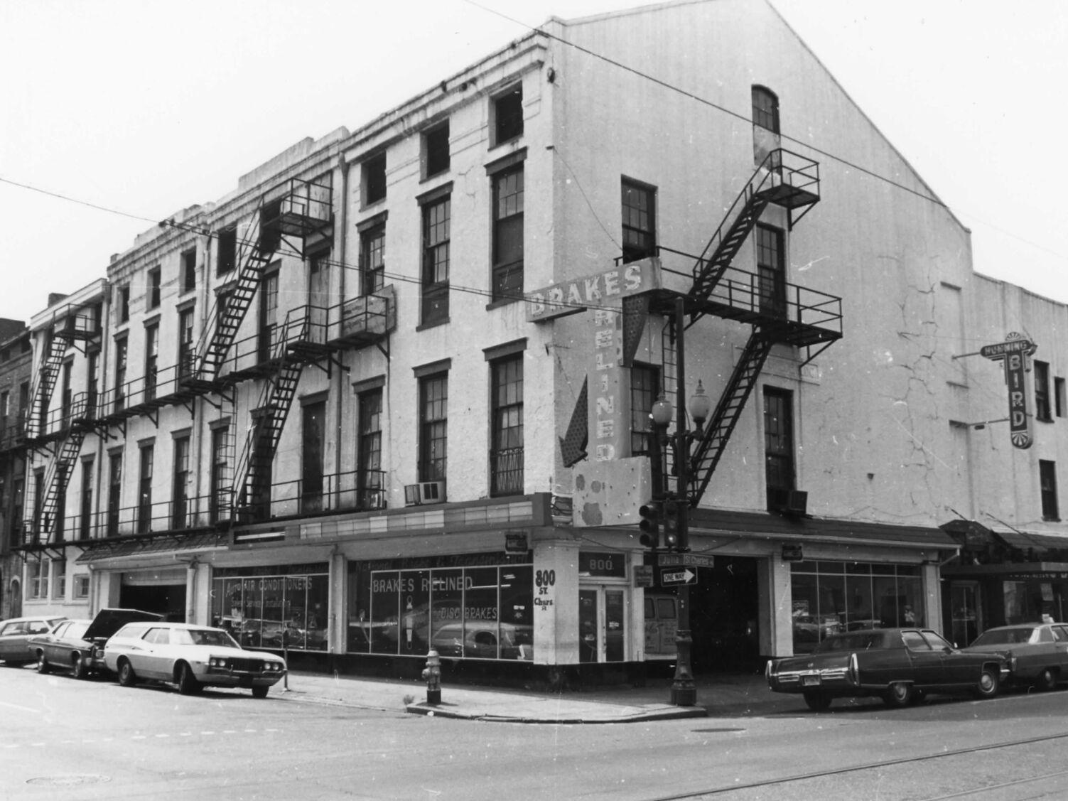 Corner warehouse building with metal fire escapes above storefronts and vintage cars parked along the street.