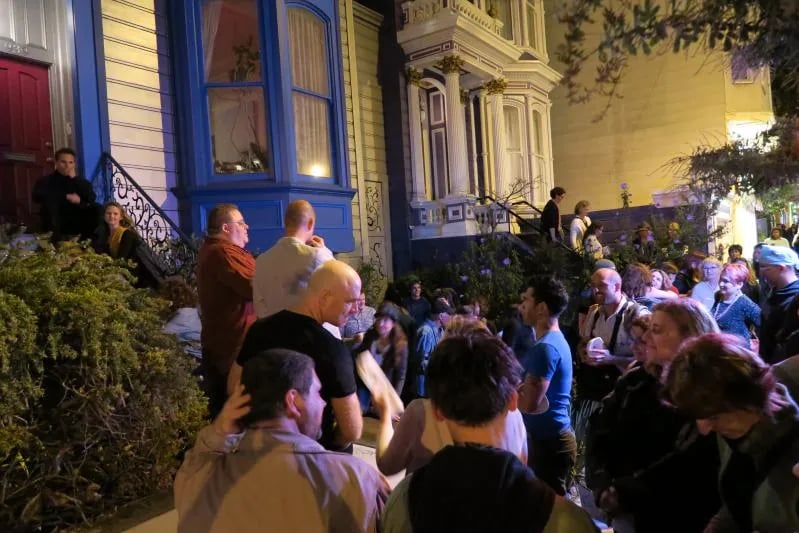 A crowd of people gathered on a sidewalk and on the front steps of a Victorian-style house at night for an outdoor reading event, with the house facade illuminated in the background.