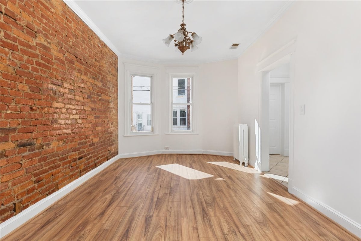 Vacant room with hardwood floors, exposed brick wall, two windows, and a chandelier.
