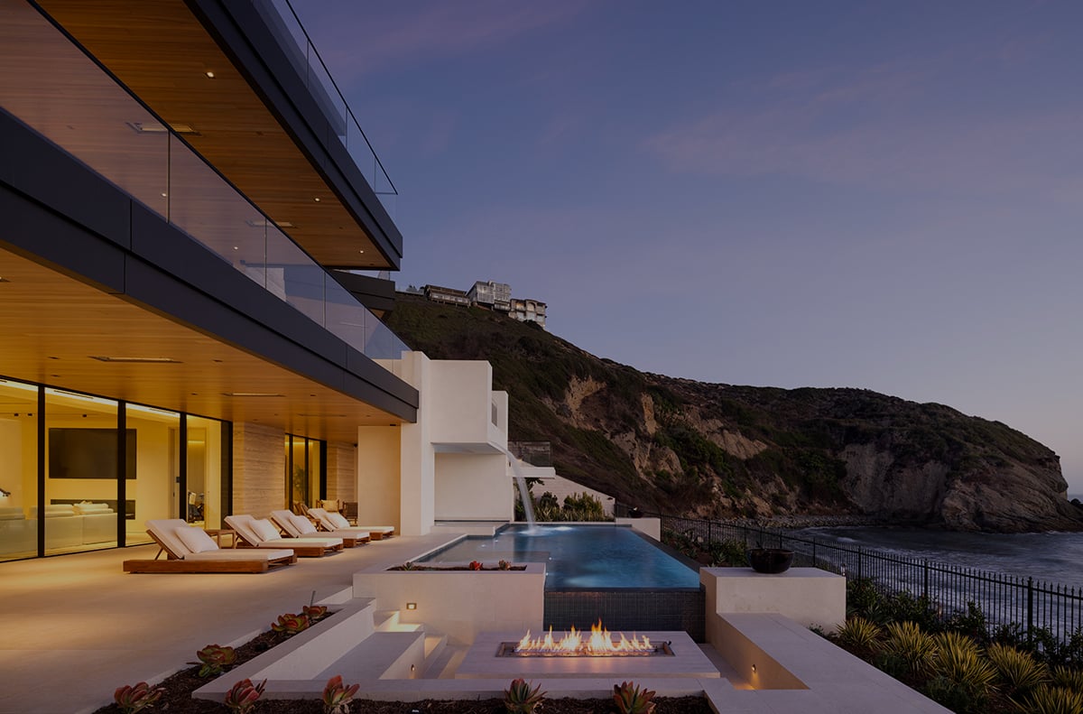 Modern coastal home with an infinity pool and fire feature overlooking the ocean at dusk.