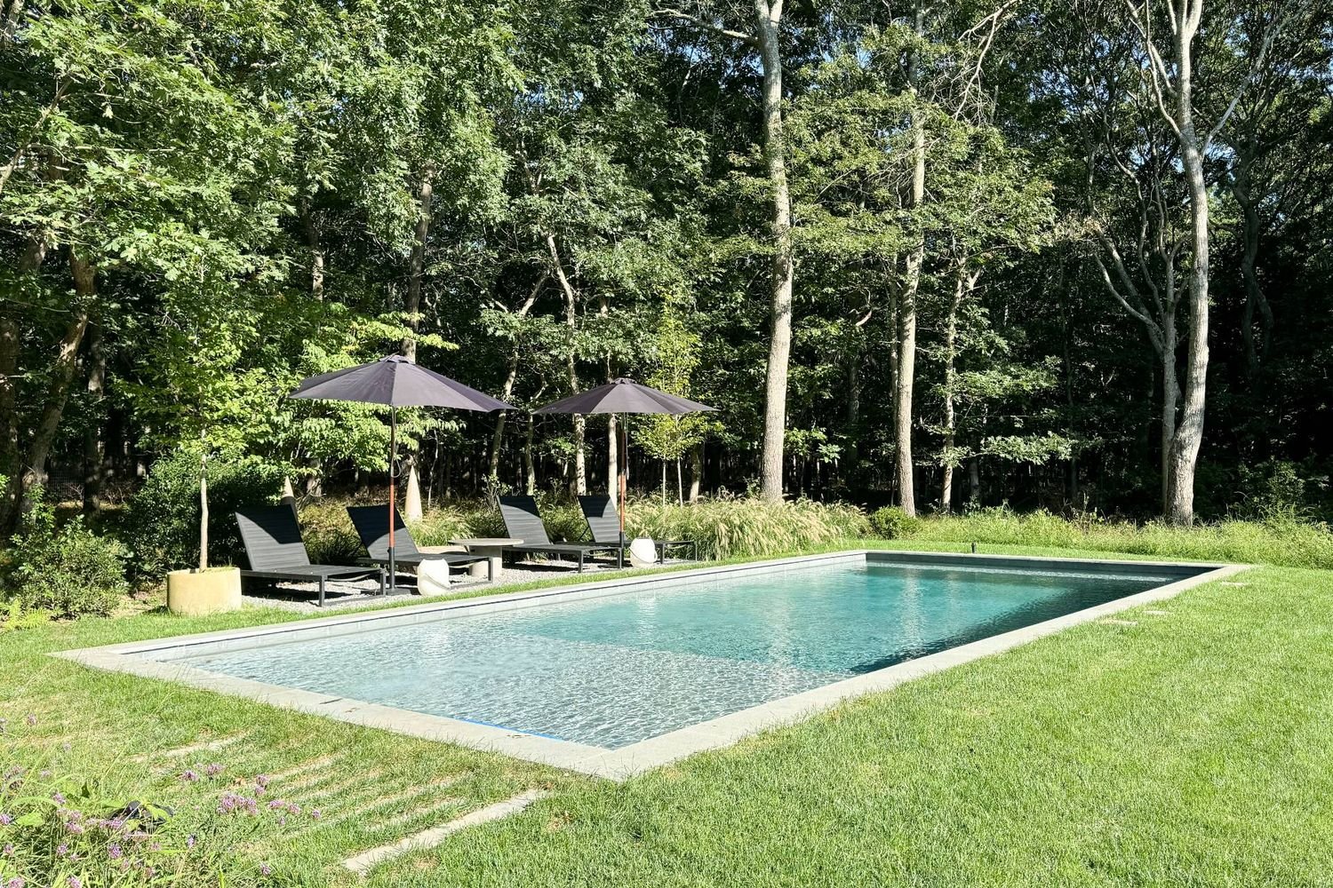 Rectangular swimming pool beside shaded lounge chairs and umbrellas, bordered by lawn and woodland.
