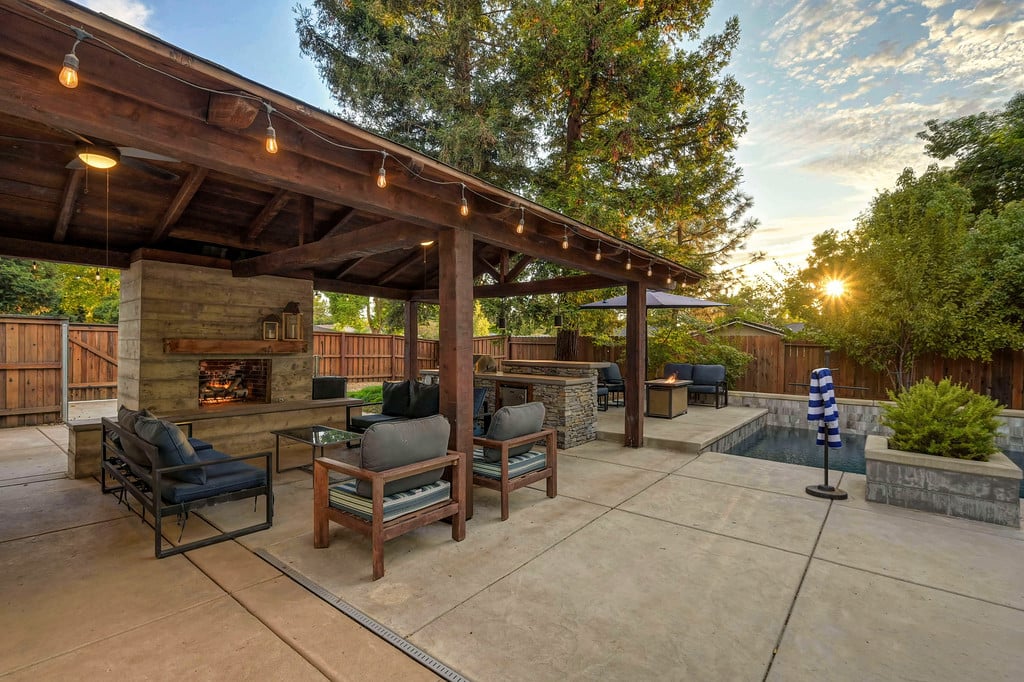 Covered patio with string lights, outdoor fireplace, lounge seating, and a pool at golden hour.