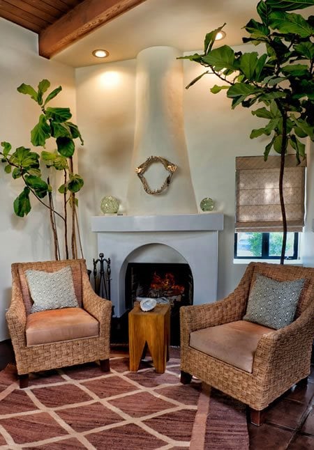 Living room with a white fireplace, wicker armchairs, leafy plants, and a wood-plank ceiling.