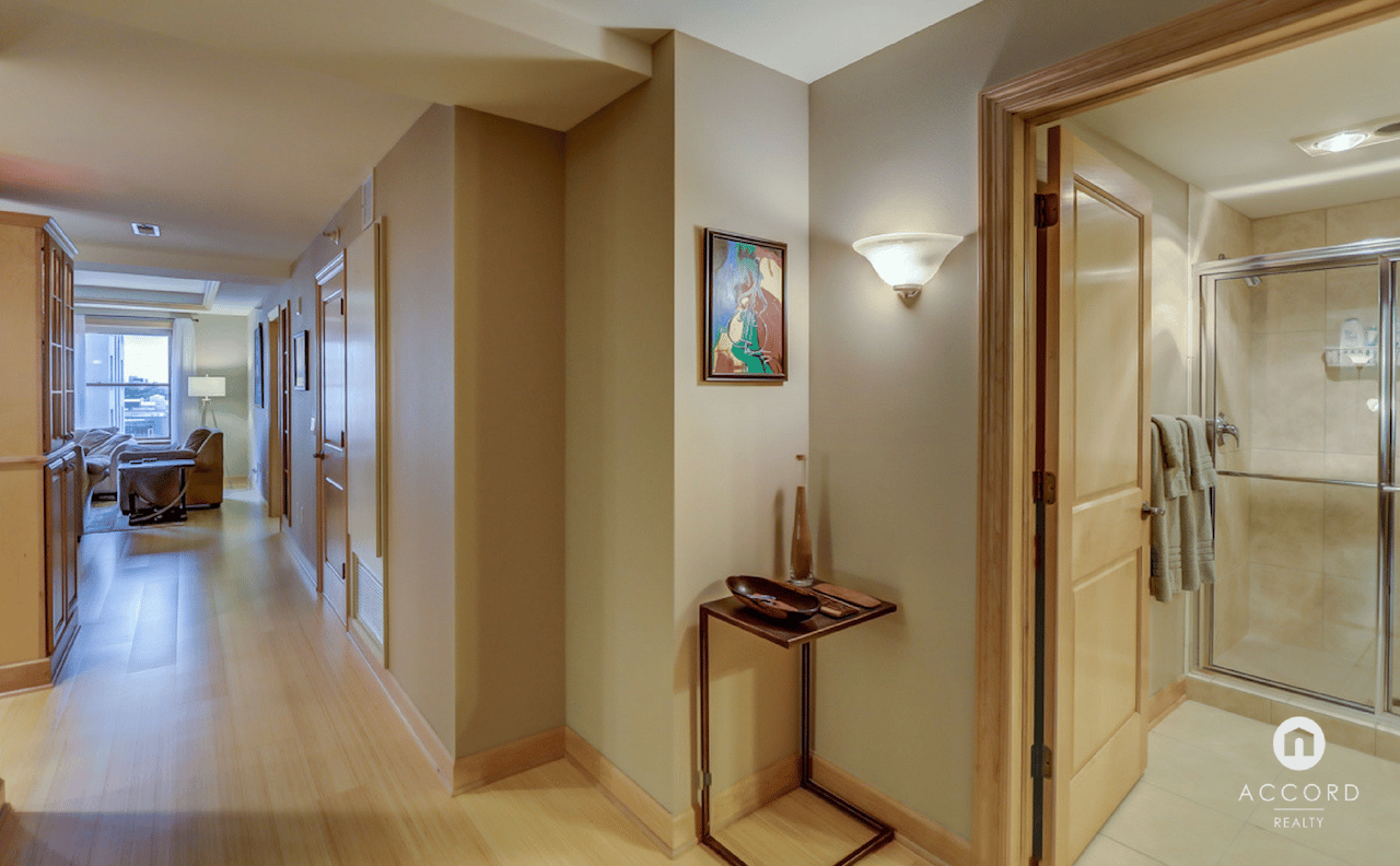 Condo hallway with wood floors, a living area by the window, and an open bathroom with glass shower.