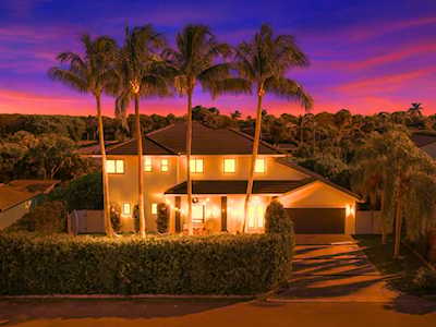 Two-story home glowing at dusk behind tall palm trees and a trimmed hedge.