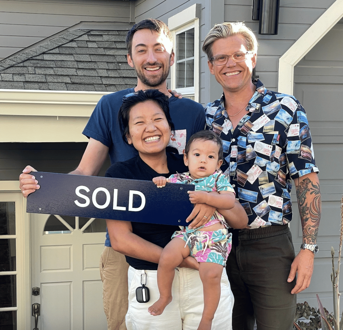 Four people pose with a SOLD sign outside a gray house.