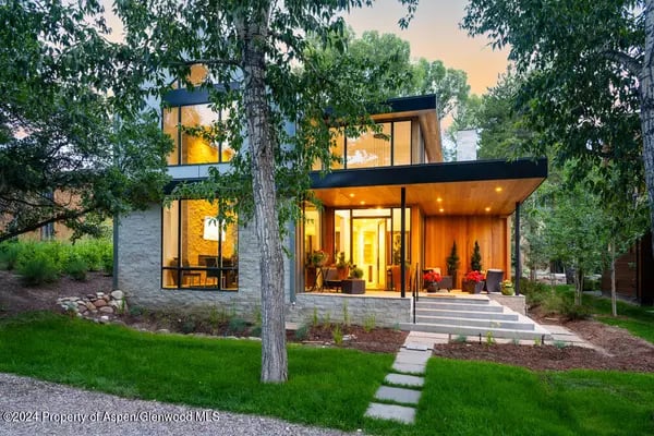 Modern glass-fronted home with a warmly lit wood entrance, mature trees, and a stone path across the lawn.