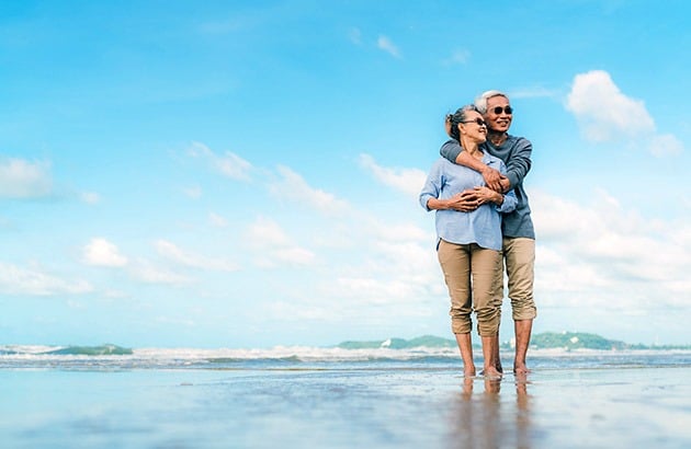 Older couple embracing in shallow beach water beneath a bright blue sky.