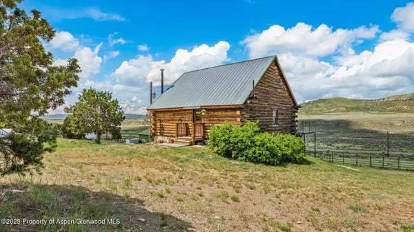 Log cabin with a metal roof in an open rural landscape, bordered by shrubs and a fence.