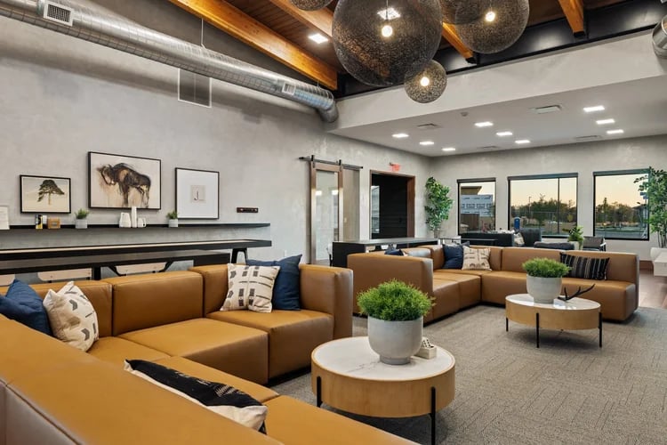 Community lounge with tan sectional sofas, exposed wooden beams, and large windows.