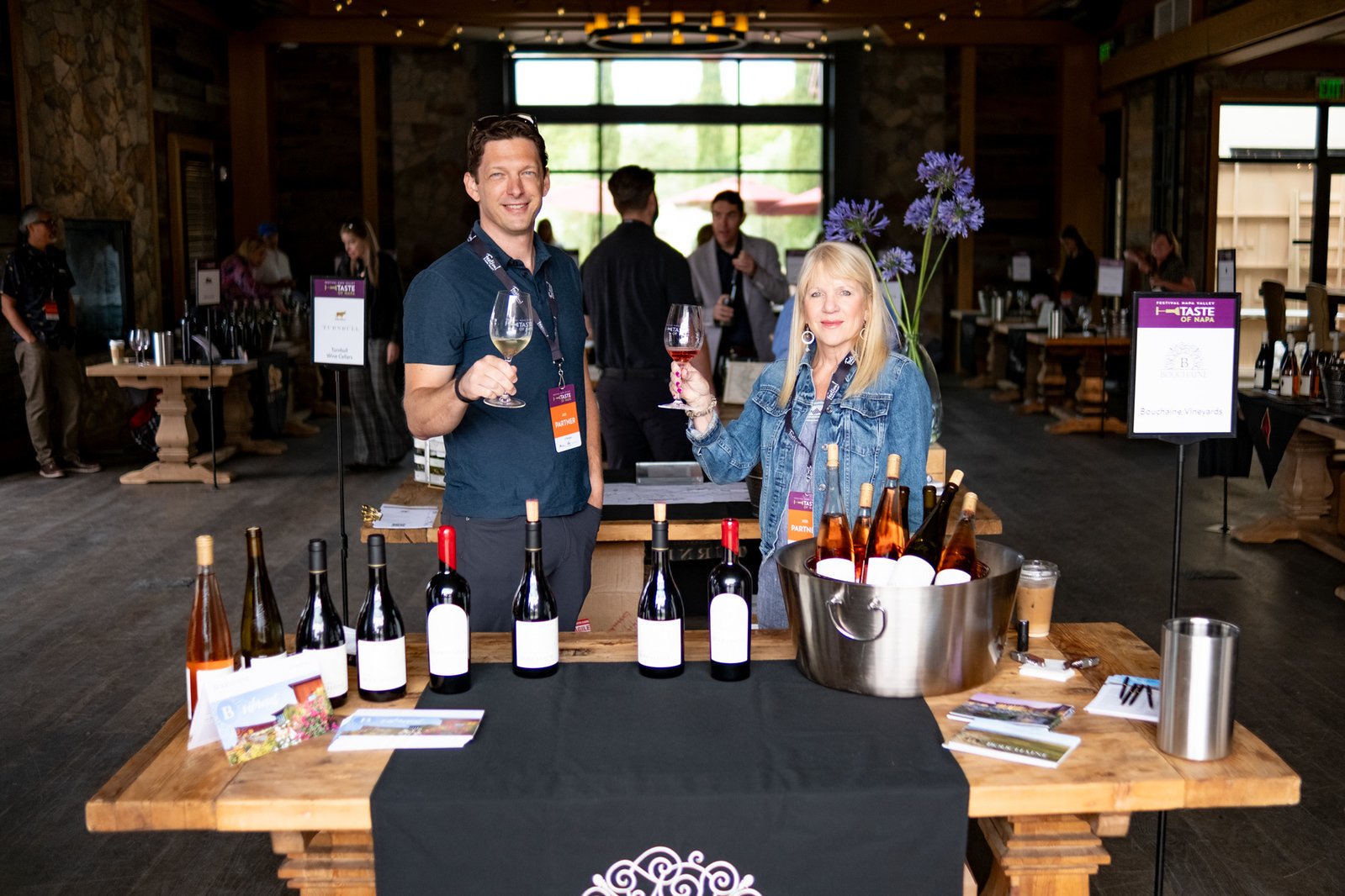Two Festival Napa Valley attendees raise wine glasses at a tasting table lined with wine bottles inside a rustic event venue.