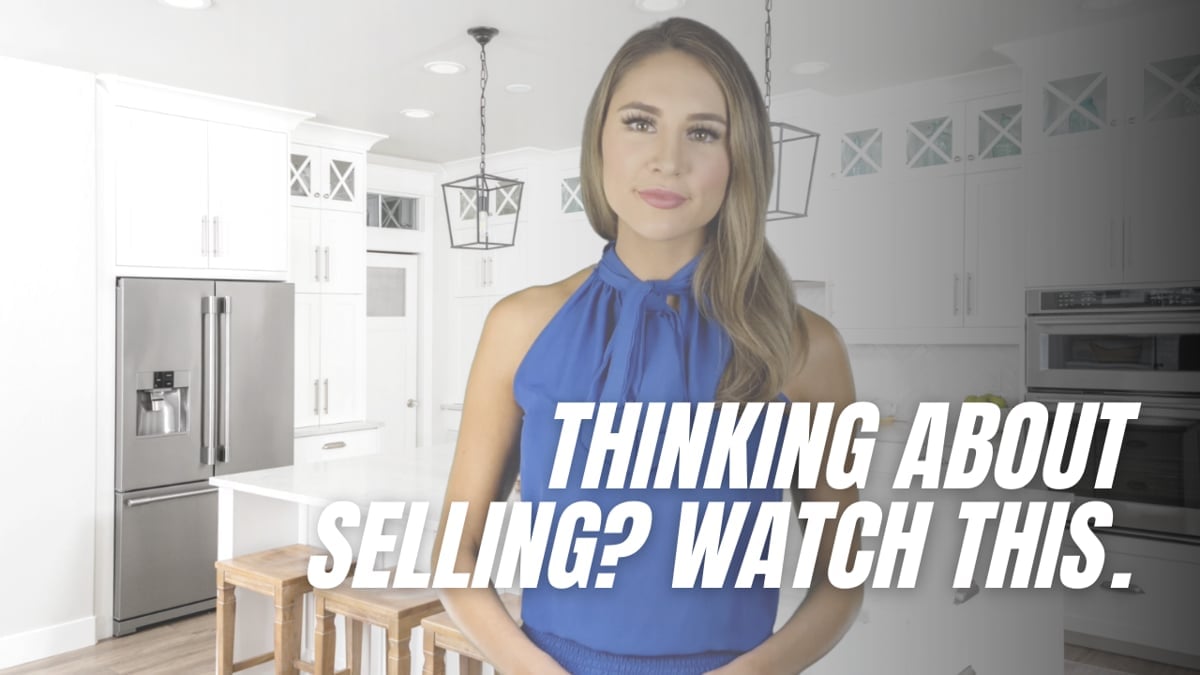 A woman in a blue blouse stands in a bright kitchen beneath the text “Thinking About Selling? Watch This.”