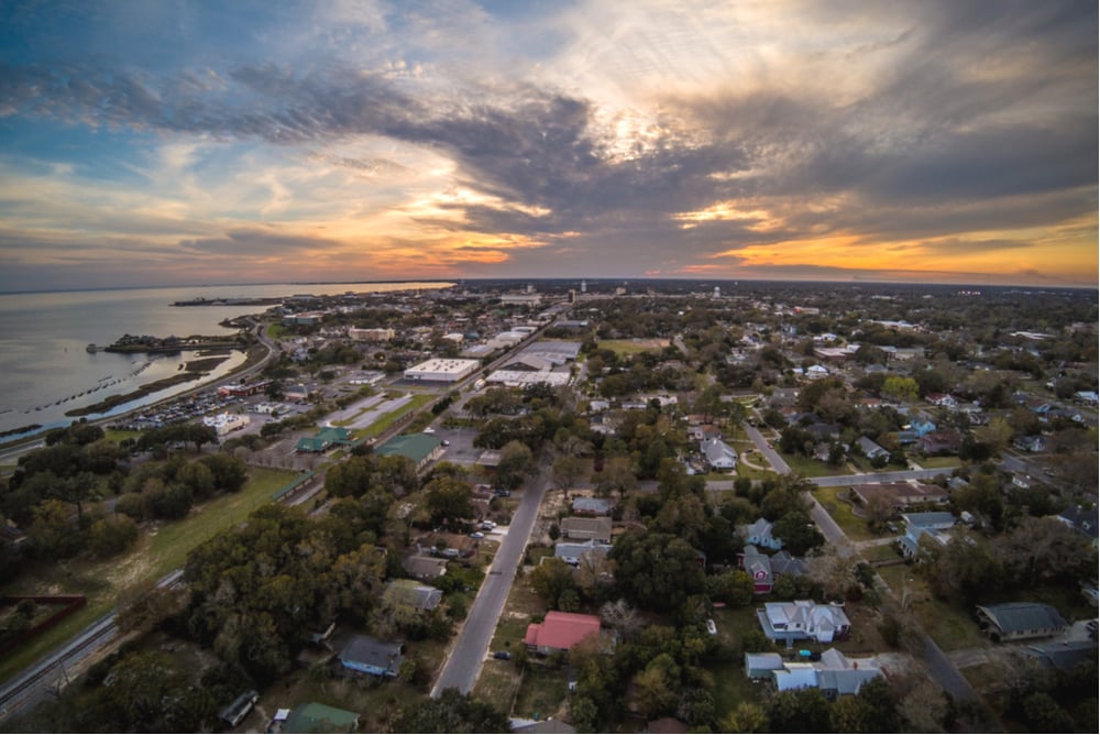 Aerial view of a coastal neighborhood at dusk, with homes, tree-lined streets and water along the shoreline.