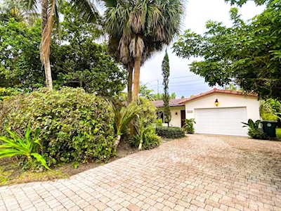 Single-story home with a two-car garage, brick-paver driveway, and mature tropical landscaping.