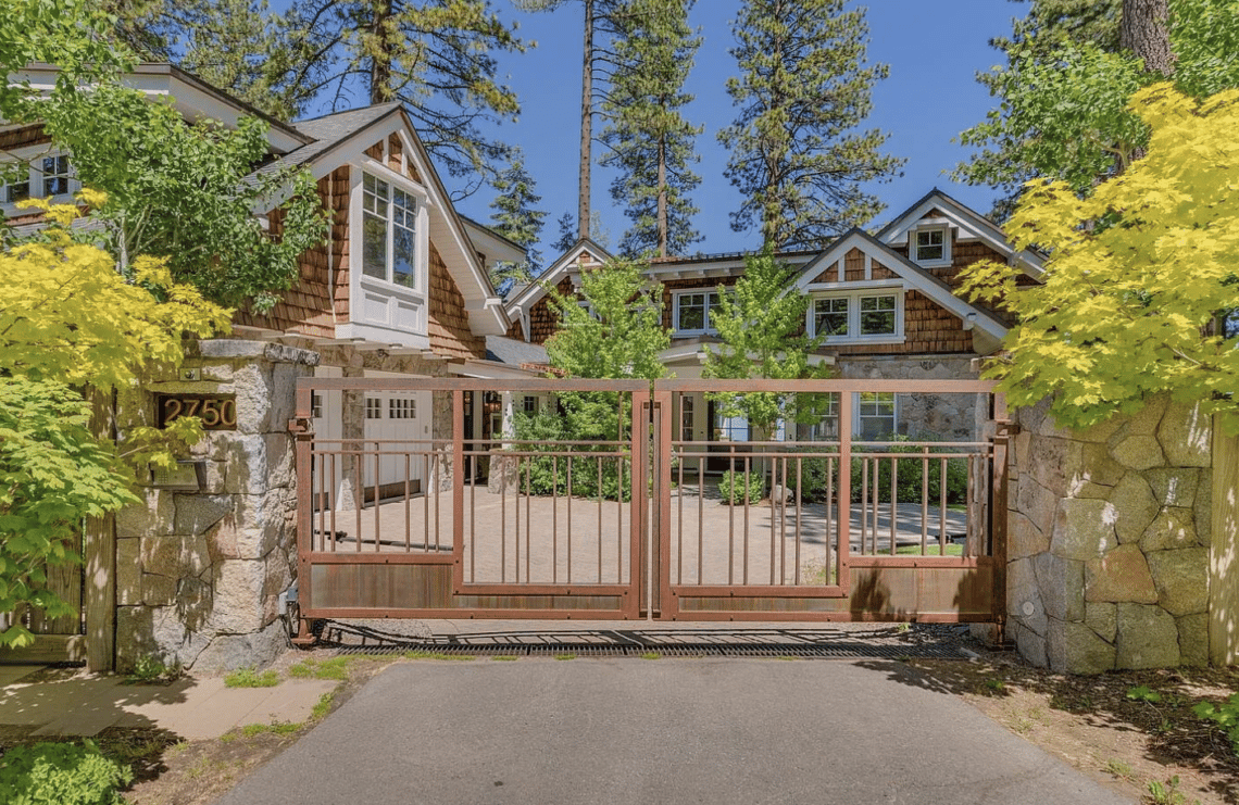 Shingle-and-stone house behind a metal gate, framed by yellow foliage and pine trees.