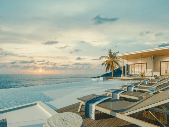 Modern beachfront villa with an infinity pool, palm tree, and lounge chairs overlooking the ocean at sunset.