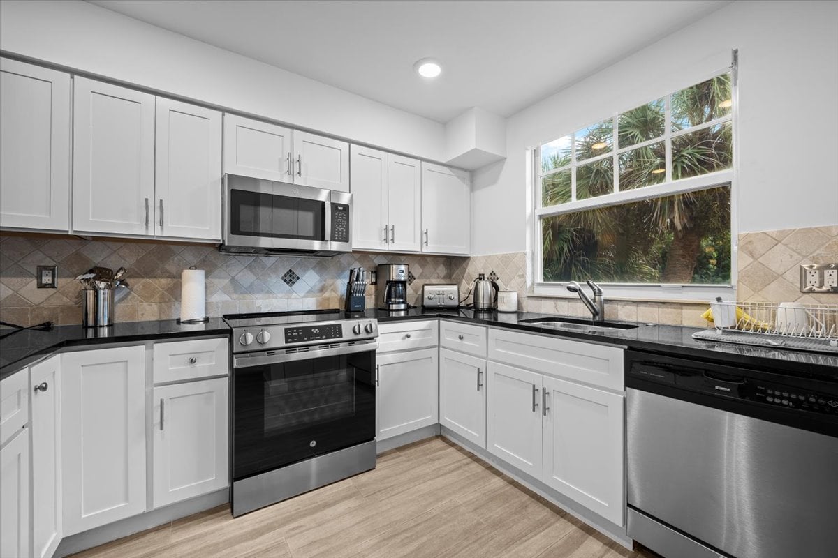 Condominium kitchen with white cabinets, stainless steel appliances, and a large window facing tropical greenery.