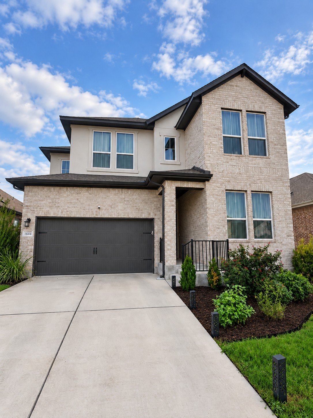 Two-story beige brick house with a dark double garage door and landscaped front garden.