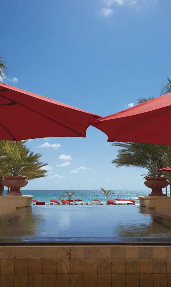 Resort pool overlooking the ocean, framed by red umbrellas and palm trees.