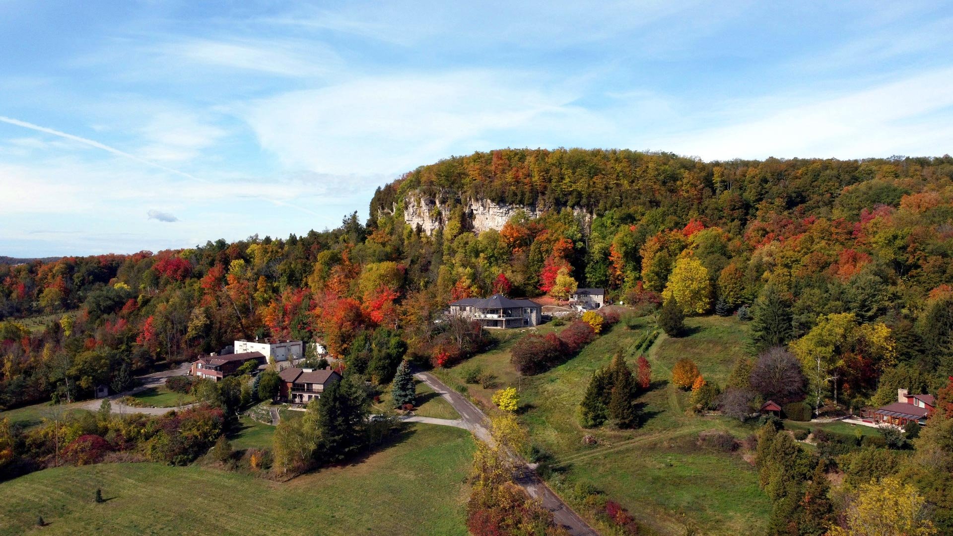 An aerial drone photograph of custom luxury homes nestled at the base of a rocky cliff along the Niagara Escarpment during autumn, surrounded by vibrant red, orange, and yellow fall foliage, showcasing the natural landscape and real estate appeal of living in Milton Ontario.
