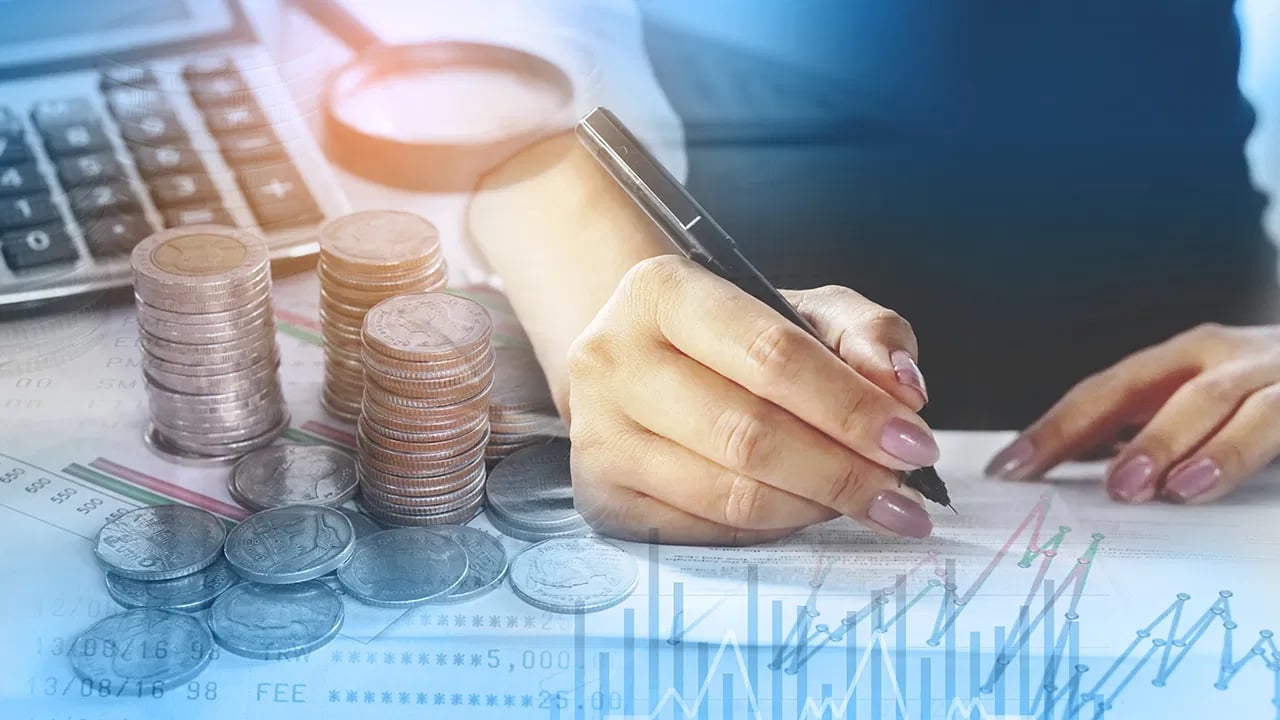 A hand writes on financial paperwork beside stacked coins and a market chart.