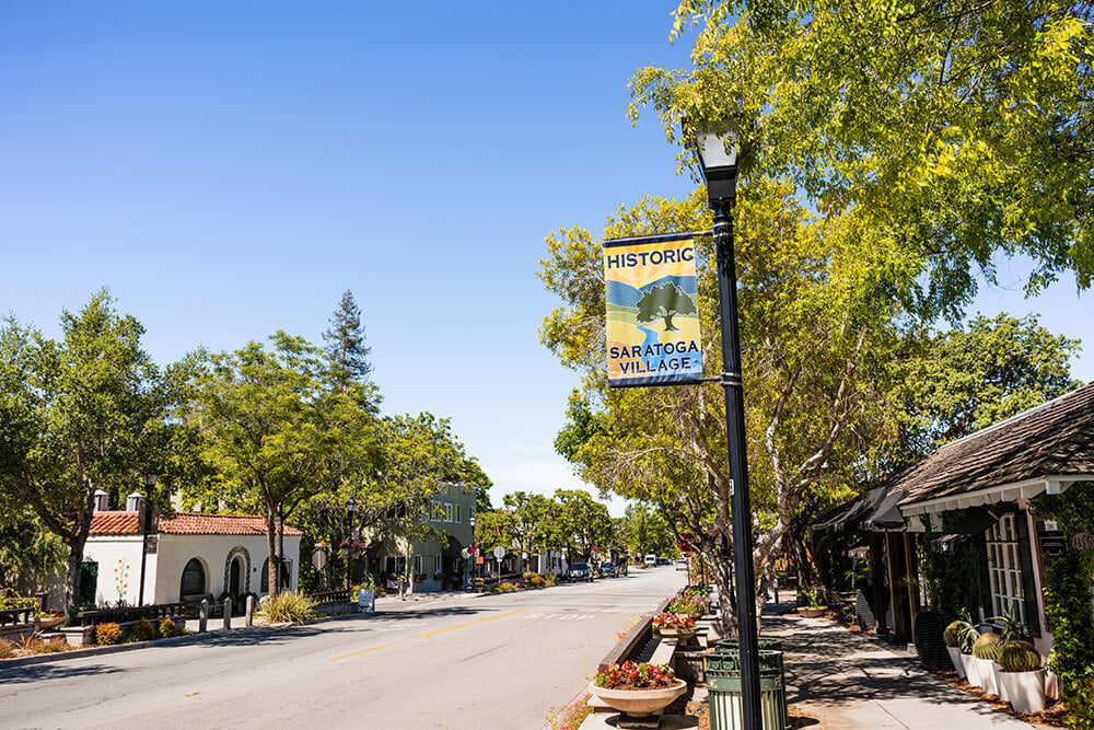 Saratoga Village street lined with leafy trees, storefronts, and flower planters beneath a Historic Saratoga Village banner.