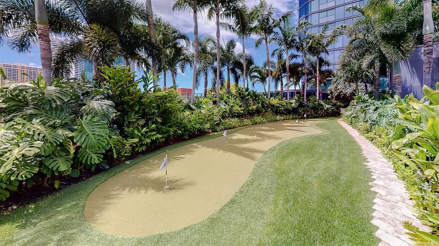 Outdoor putting green with several flagsticks, tropical palms, and a stone path beside a glass building.