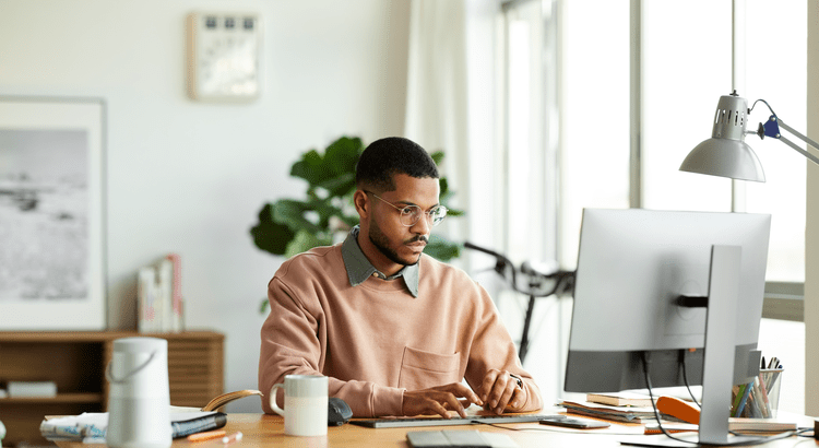 A focused man in glasses types on a computer in a bright home office with a plant, bookshelf, and window. The setting is calm and productive.