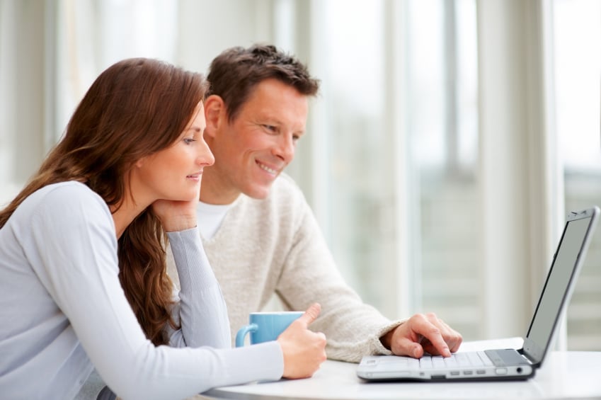 Couple look at a laptop together; the woman holds a blue mug as the man points at the screen.