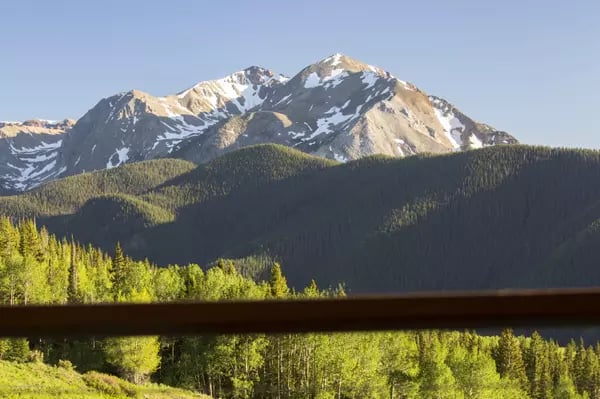 Snow-dusted mountain peaks above forested slopes and sunlit trees, with a wooden railing in the foreground.