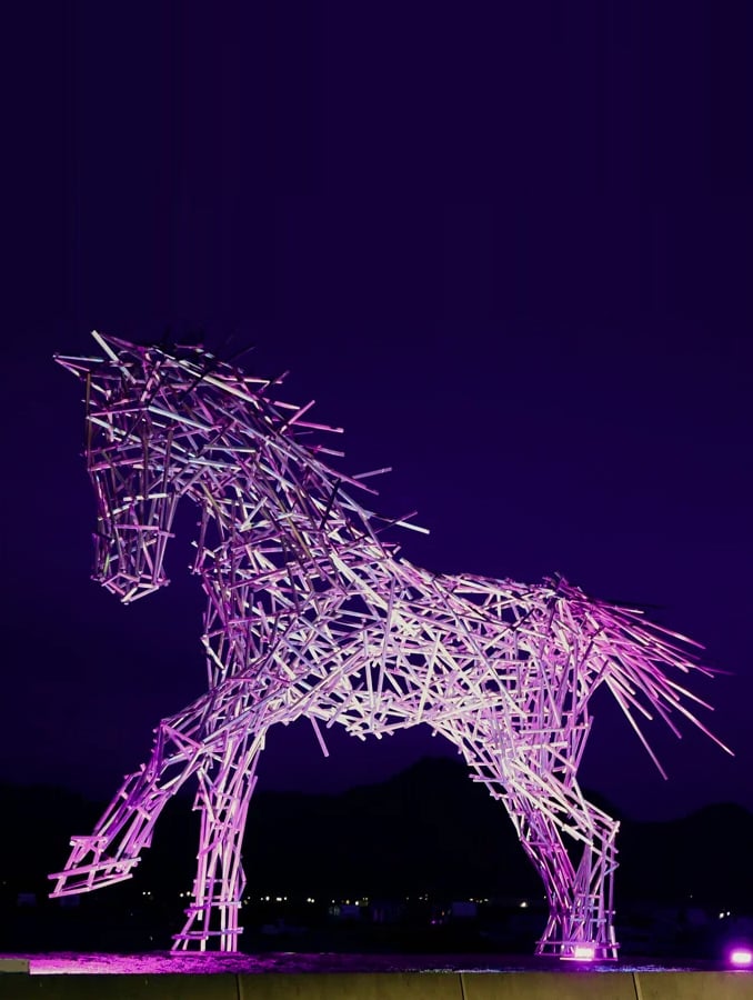 Glowing pink-and-white metal horse sculpture standing against dark mountains at night.
